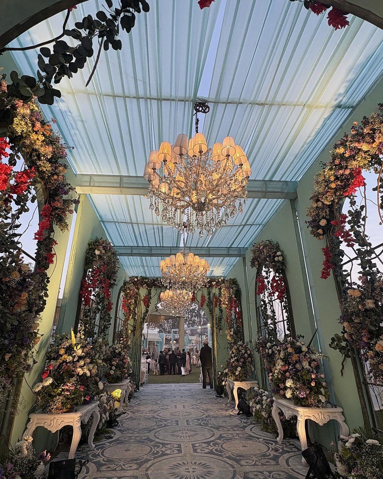 A floral tunnel on a patterned floor under two chandeliers, in daylight