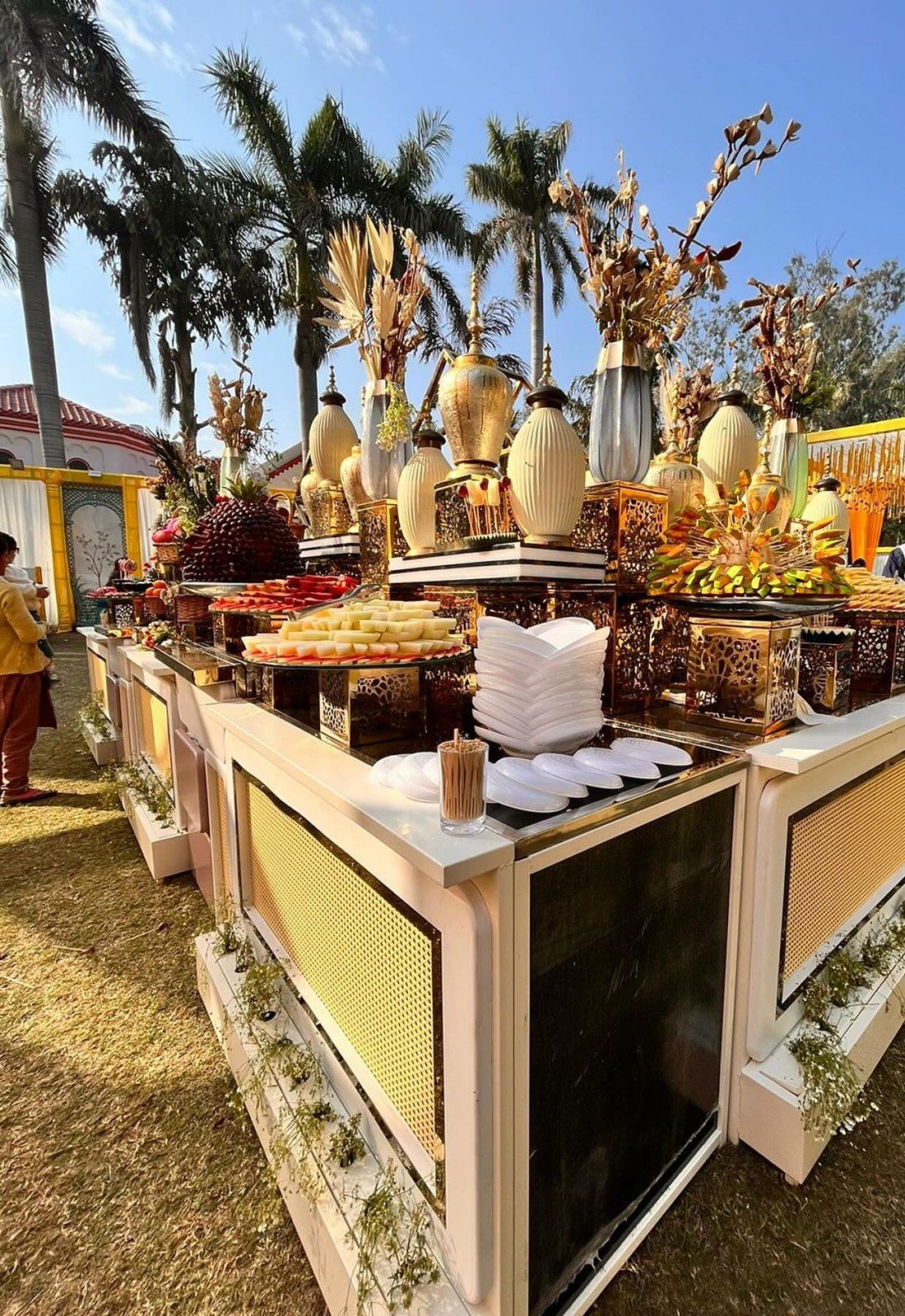A buffet counter on a lawn under palm trees, with brass urns and stacked plates