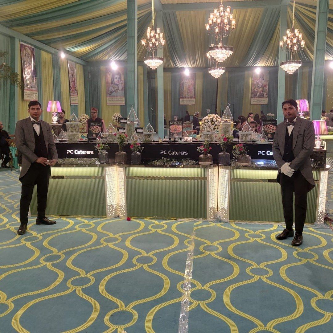Two suited staff beside a counter on a patterned carpet under chandeliers
