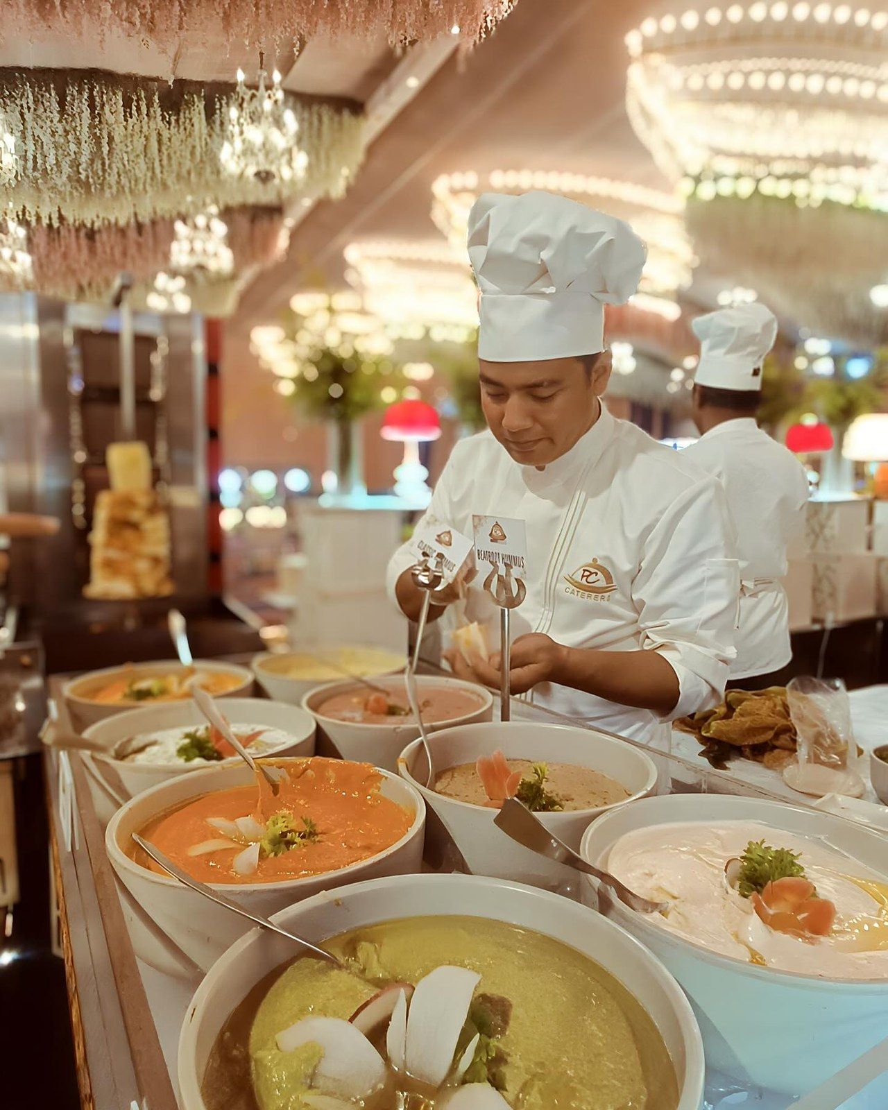 A chef in a white toque serving from bowls of soup under chandeliers