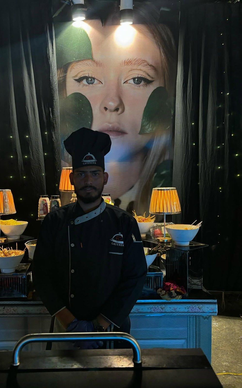 A server in a dark chef coat holding a lamp-lit tray, a portrait print behind